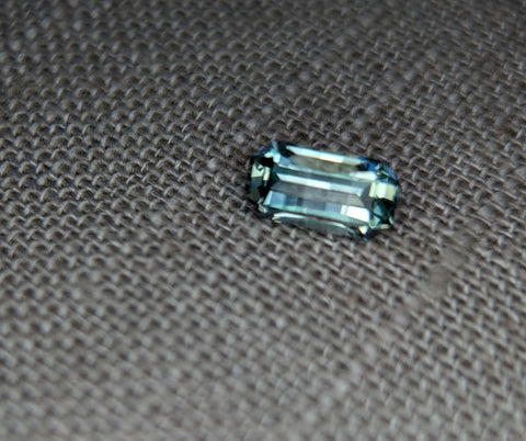 A single aqua green/blue Montana sapphire gemstone with a custom emerald cut, placed on a textured grey background.
